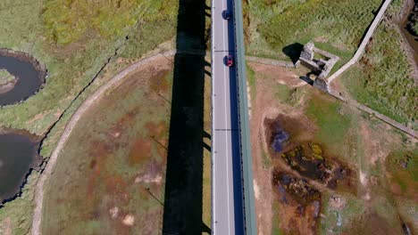 Cars-circulating-over-the-bridge-over-the-Ulla-River-in-drought-with-the-nature-that-emerges-from-the-bed,-sunny-afternoon,-rolling-drone-shot-up-zenithal