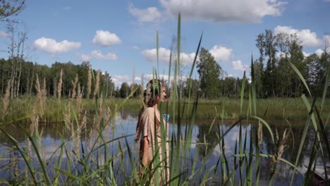 A-woman-is-walking-by-the-pond-as-the-camera-is-following-her-with-tall-grasses-in-the-foreground,-natural-colour,-slow-motion