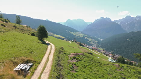 Drone-video-flying-low-over-mountain-path-and-rising-vertically-towards-Dolomites-mountain-range-in-summer-flying-over-historical-hillside-town-surrounded-by-forests-and-farmland