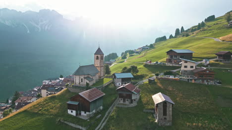 Drone-video-flying-over-historical-hillside-town-surrounded-by-farms-in-the-Dolomites-Italy-passing-medieval-catholic-church-with-mountain-range-in-the-background-at-sunset-golden-hour-in-summer