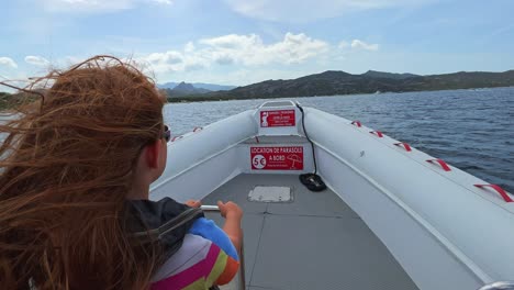 Back-view-of-long-hair-redhead-child-with-sunglasses-on-motorboat-sailing-over-north-Corsica-sea-water-for-tour-toward-Saleccia-famous-beach