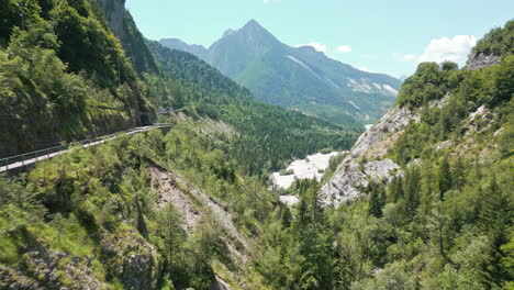 Drone-video-flying-over-forest-to-mountain-road-bridge-panning-up-to-reveal-dry-valley-in-the-Italian-Dolomites-surrounded-by-mountain-roads-and-cliffs-and-small-lake-below