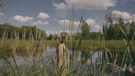 A-woman-is-walking-by-the-pond-as-the-camera-is-following-her-with-tall-grasses-in-the-foreground,-colour-graded,-slow-motion