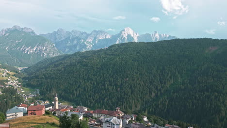 Drone-video-flying-by-historical-hillside-catholic-church-in-old-town-surrounded-by-farms-in-the-Dolomites-Italy-with-mountain-range-in-the-background-at-sunset-golden-hour-in-summer