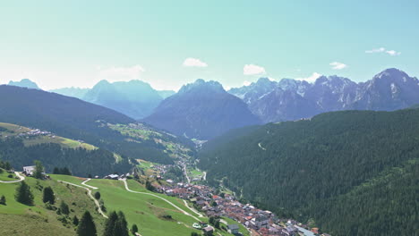 Drone-video-flying-towards-Dolomites-mountain-range-in-summer-flying-over-historical-hillside-town-surrounded-by-forests-and-farmland