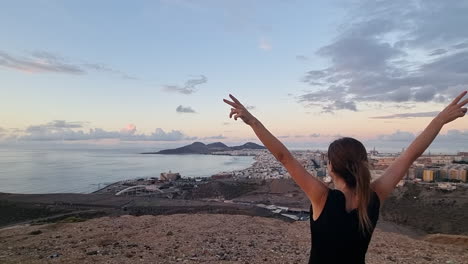 Woman-admires-with-raised-arms-and-from-a-viewpoint,-at-sunset,-Las-Canteras-beach-and-the-Alfredo-Kraus-auditorium