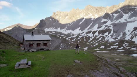 Aerial-drone-footage-pflying-over-a-man-standing-next-to-a-Swiss-alpine-hut-in-a-dramatic-mountain-landscape-with-residual-patches-of-snow-and-alpine-meadows-in-Switzerland