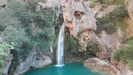 Aerial-elevating-shot-over-a-spectacular-waterfall,-skull-shape-rock,-as-the-power-of-nature-formed-a-clear-green-lake-in-the-mountains