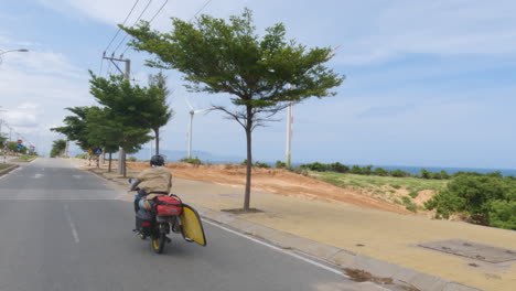 Man-riding-motorbike-carrying-surfboard-in-Indonesia,-follow-back-view