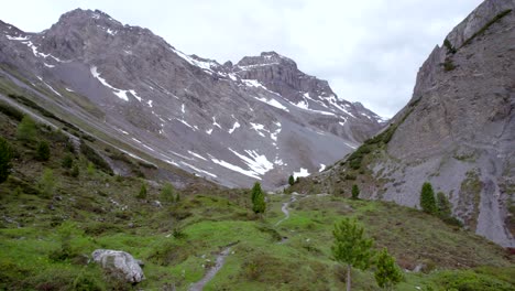 Aerial-drone-footage-rolling-through-a-glacial-mountain-landscape-with-patches-of-snow,-isolated-trees-a-remote-alpine-hiking-trail-in-Switzerland