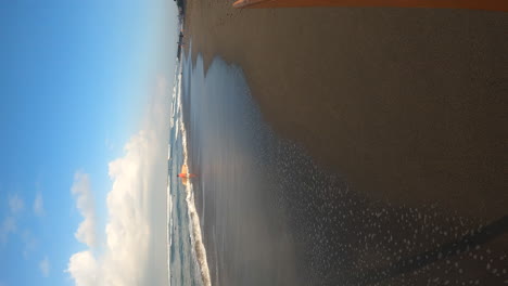 Person-carrying-surfboard-and-walking-on-sandy-beach-in-Bali,-POV-vertical-view
