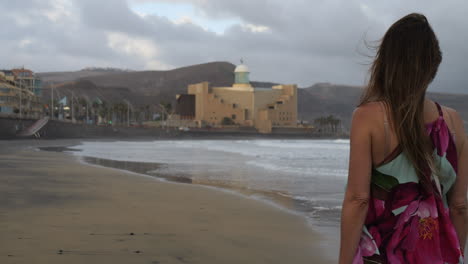 Cinematic-shot-at-sunset-of-a-woman-walking-along-Las-Canteras-beach-and-seeing-the-Alfredo-Kraus-Auditorium-in-the-background