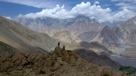Two-people-standing-on-peak-at-Passu-Cones-Pakistan,-cinematic-wide-revealing-aerial-shot