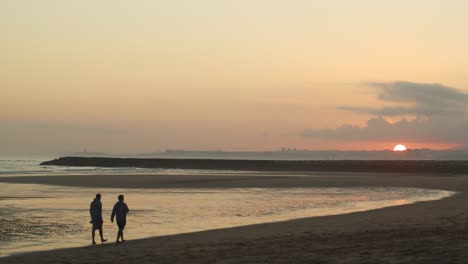 Two-male-friends-walking-on-beach-at-sunset,-wide-shot