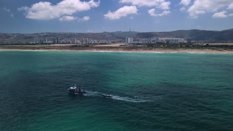 Drones-Rodeando-Barcos-De-Pesca-Lanzando-Redes-Mientras-Revelan-La-Ciudad-De-Hof-Hacarmel-En-El-Fondo-Con-La-Playa-De-Haifa-Y-El-Cielo-Brillante