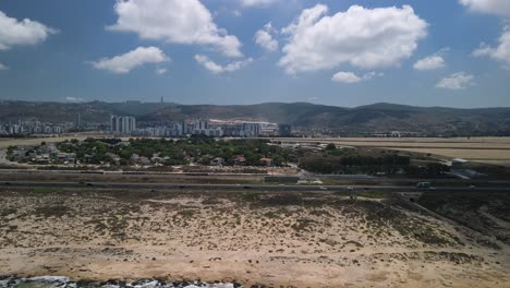 Drone-Moviéndose-Desde-La-Playa-De-Haifa-A-La-Ciudad-Como-Un-Cruce-De-Carreteras-Ocupado-Con-Vehículos,-Con-Un-Agradable-Cielo-Azul-Con-Montañas-Distantes