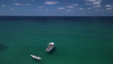 Die-Drohne-Bewegt-Sich-An-Einem-Verankerten-Fischerboot-Vorbei-Und-Fährt-Auf-Einen-Leuchtenden-Horizont-Zu,-Der-Das-Aufeinandertreffen-Des-Türkisfarbenen-Ozeans-Und-Des-Blauen-Himmels-Zeigt