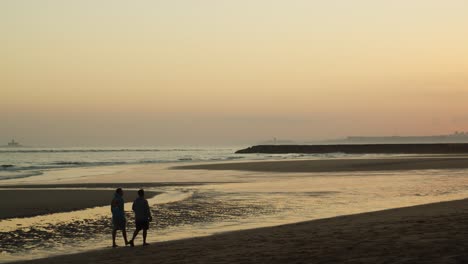 Two-male-friends-walking-on-beach-at-sunset,-handheld-wide-shot