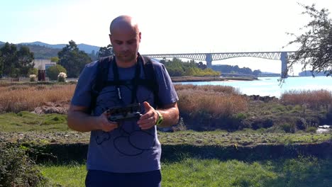 40-year-old-male-drone-pilot-making-adjustments-and-inspecting-looking-straight-at-the-camera-with-a-bridge-and-the-river-behind-it,-sunny-summer-afternoon,-frontal-shot
