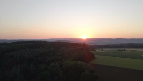 Bird's-eye-view-at-sunset-of-meadows-and-cornfields-in-a-valley-surrounded-by-forests