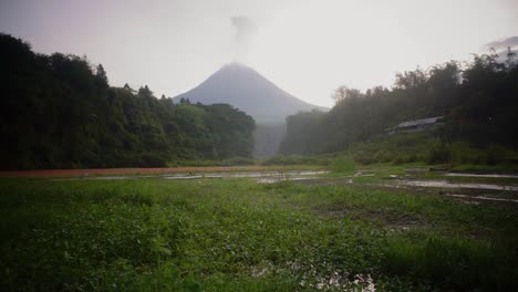 Bergfluss-Bedeckt-Von-Grünem-Gras-Mit-Vulkan,-Der-Rauch-Ausstößt---Wunderschöne-Naturaufnahmen-Im-Dschungel