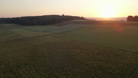 Backward-drone-flight-at-sunset-over-an-agricultural-field-surrounded-by-forests-and-hills