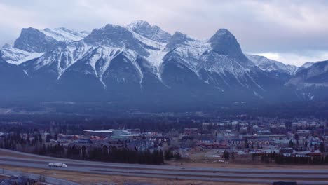 18-wheeler-truck-and-cars-ride-highway-in-foreground-with-three-sisters-mountain-range-in-Canmore-Alberta-in-Canada,-sweeping-dynamic-drone-shot