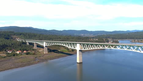 Railway-bridge-over-the-ulla-river,-the-village,-industrial-buildings-and-the-shore,-the-sunny-blue-sky-orizonte,-drone-shot-traveling-diagonally-to-the-left,-Catoira,-Galicia,-Spain