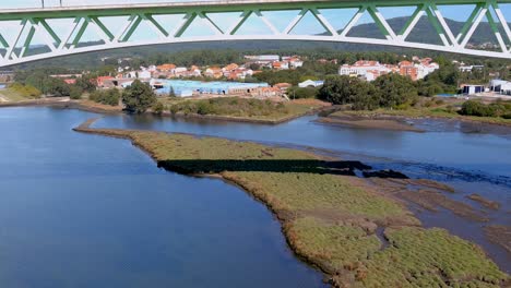 Below-the-railway-bridge-with-power-lines-over-the-Ulla-River-with-low-water-level,-industrial-buildings-behind,-drone-shooting-traveling-forward,-below,-Catoira,-Galicia,-Spain