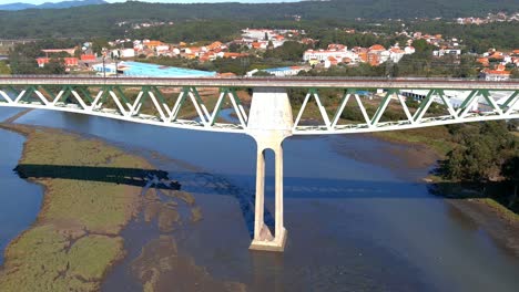 Railway-bridge-with-power-lines-over-the-ulla-river-with-low-water-level,-seagulls-flying-low,-industrial-buildings-behind,-shot-of-rolling-overhead-drone-traveling-forward,-Catoira,-Galicia,-Spain
