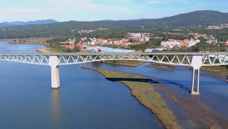 Railway-bridge-over-the-ulla-river,-the-village,-industrial-buildings-and-the-forested-mountains-in-the-sunny-blue-sky-orizonte,-drone-shot-traveling-forward-down,-Catoira,-Galicia,-Spain