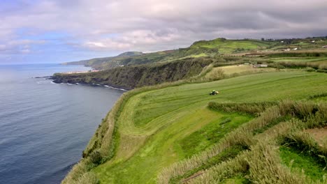 Tractor-harvesting-green-field-on-sea-coast-cliff,-Azores,-flyover
