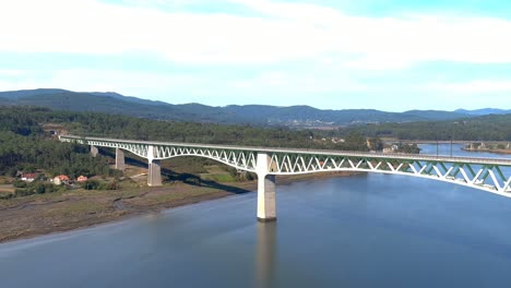 Railway-bridge-over-the-ulla-river,-the-village,-industrial-buildings-and-the-shore,-the-sunny-blue-sky-orizonte,-drone-shot-traveling-backwards-diagonally,-Catoira,-Galicia,-Spain
