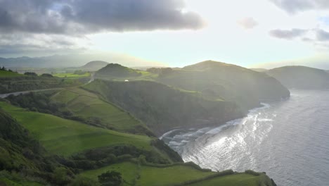 Clouds-breaking-over-sunlit-grassy-coast-cliffs,-Azores,-aerial-view