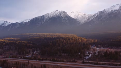 Canmore-Alberta-mountain-range-Canadian-rockies-background-aerial-sunrise-drone-shot,-crane-up-highway-with-cars-driving-by,-big-condo-complex-in-foreground