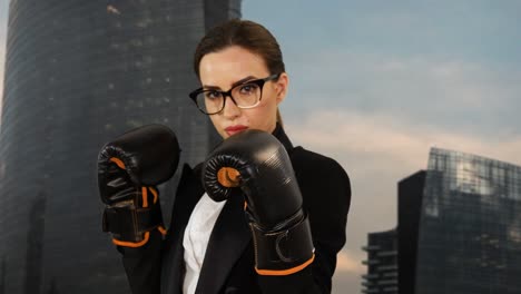 Businesswoman-fight-against-stress-due-to-hard-work-by-wearing-boxing-gloves-and-punching-toward-camera-with-skyscrapers-in-background-and-moving-clouds