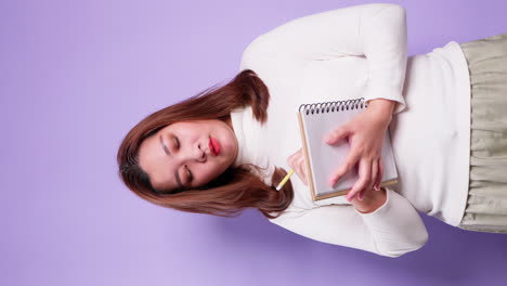 Vertical-of-Asian-secretary-businesswoman-holding-a-notebook-and-thinking-with-list-inspiration-strategy-for-success-on-purple-background
