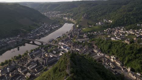 Wide-angle-aerial-dive-down-a-steep-mountain-ridge-towards-the-old-town-and-the-moselle-river-in-Cochem,-Germany-in-soft-morning-light