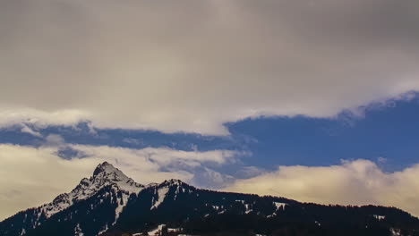 Time-Lapse-of-snow-capped-mountain-peak,-grey-clouds-moving-fast-with-blue-sky