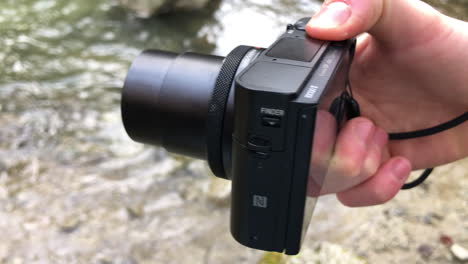 A-compact-camera-unfolds-its-lens,-seen-from-above-on-the-edge-of-a-Swiss-river,-technology