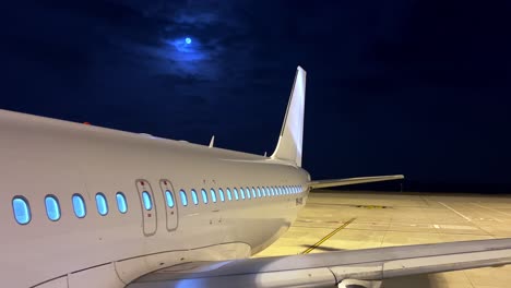 Nachtansicht-Des-Geparkten-Flugzeugs-Auf-Dem-Vorfeld-Des-Flughafens-Mit-Weißem-Körper,-Der-Rumpfflügelmotor-Und-Heck-Mit-Mond-Im-Hintergrund-Zeigt