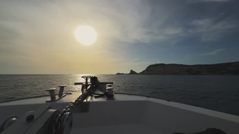 Point-of-view-of-sailboat-bow-navigating-toward-Corsica-island-cliffs-and-Capo-Pertusato-lighthouse-in-France-with-sun-in-background-at-sunset,-Slow-motion