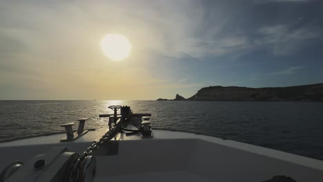 Point-of-view-of-sailboat-bow-navigating-toward-Corsica-island-cliffs-and-Capo-Pertusato-lighthouse-in-France-with-sun-in-background-at-sunset,-50fps-horizontal-footage