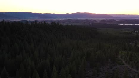 An-aerial-shot-of-over-a-dark-forest-with-the-sun-behind-the-distant-pink-and-blue-lit-mountains