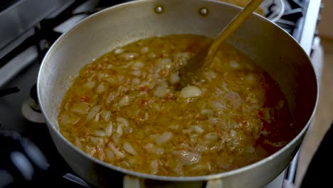 Chopped-Onions-Being-Stirred-In-Curry-Sauce-In-Large-Steel-Pot-With-Wooden-Spatula