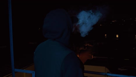 Medium-dramatic-shot-of-a-person-in-a-black-hoodie-standing-in-a-dark-room-overlooking-the-city-at-night-smoking-and-blowing-out-the-smoke-from-behind,-slow-motion