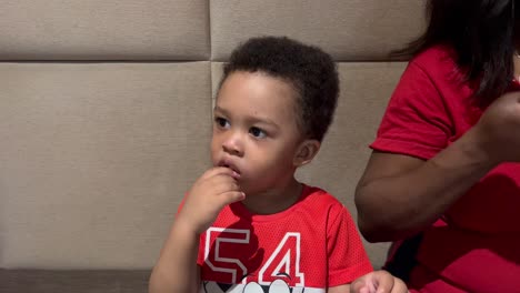 Lovely-two-year-old-black-kid-eats-italian-grissini-for-the-first-time,-seated-inside-a-restaurant-next-to-mum
