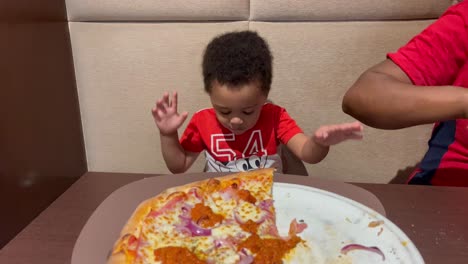 Exotic-and-adorable-two-year-old-kid-eating-pizza-for-the-first-time-inside-a-restaurant,-seated-next-to-mum,-both-of-them-wearing-red-shirts