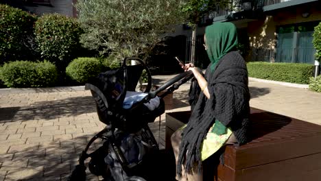 Eine-Junge-Asiatische-Muslimische-Mutter-überprüft-Ihr-Telefon,-Während-Sie-Sich-An-Einem-Sonnigen-Tag-Mit-Ihrem-Neugeborenen-In-Einem-Kinderwagen-Hinsetzt