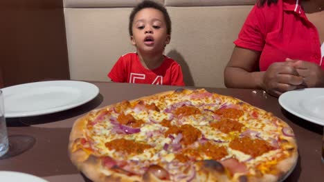 Adorable-two-year-old-child-impatient-to-eat-his-first-pizza-inside-a-restaurant,-seated-next-to-mum